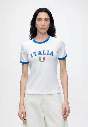 Mujer con cabello largo y oscuro que lleva una camiseta blanca con ribete azul, texto "ITALIA" y bandera italiana, combinada con pantalones blancos, de pie contra un fondo blanco.