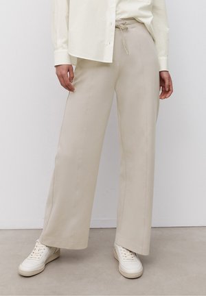 Person iført beige bukser med brede ben og bindebånd i taljen, hvid skjorte med knapper og hvide sneakers, stående på en grå gulv.