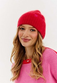 Červená pletená čiapka s mierne voľným strihom a rebrovanou štruktúrou, s malou ružovou značkovou etiketu. Modelka má pod ňou ružový sveter.