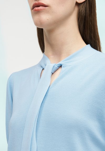 Femme aux longs cheveux bruns portant un haut bleu clair avec un nœud au niveau de l'encolure, vue du menton à la poitrine.