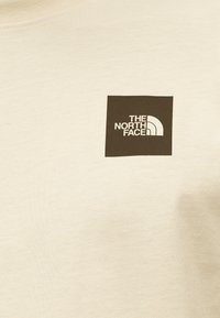 Μπεζ φούτερ κατασκευασμένο από μαλακό ύφασμα με μαύρο τετράγωνο λογότυπο που φέρει το κείμενο "THE NORTH FACE" και λευκό τόξο στο γωνία.