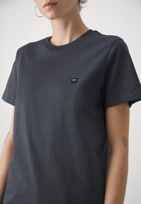 Donkergrijze katoenen t-shirt met korte mouwen, ronde halslijn en een klein zwart logopatchje op de linkerkant van de borst. Glad textuur.