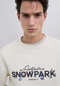 Sudadera crema con el texto "Sculptor SNOWPARK APRÈS SKI" y bordado colorido con temática de esquí. Textura suave, cuello redondo, ajuste relajado.
