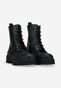 Botas de combate de cuero negro con cordones, suelas de goma gruesas y cremalleras laterales, colocadas una al lado de la otra sobre un fondo blanco.