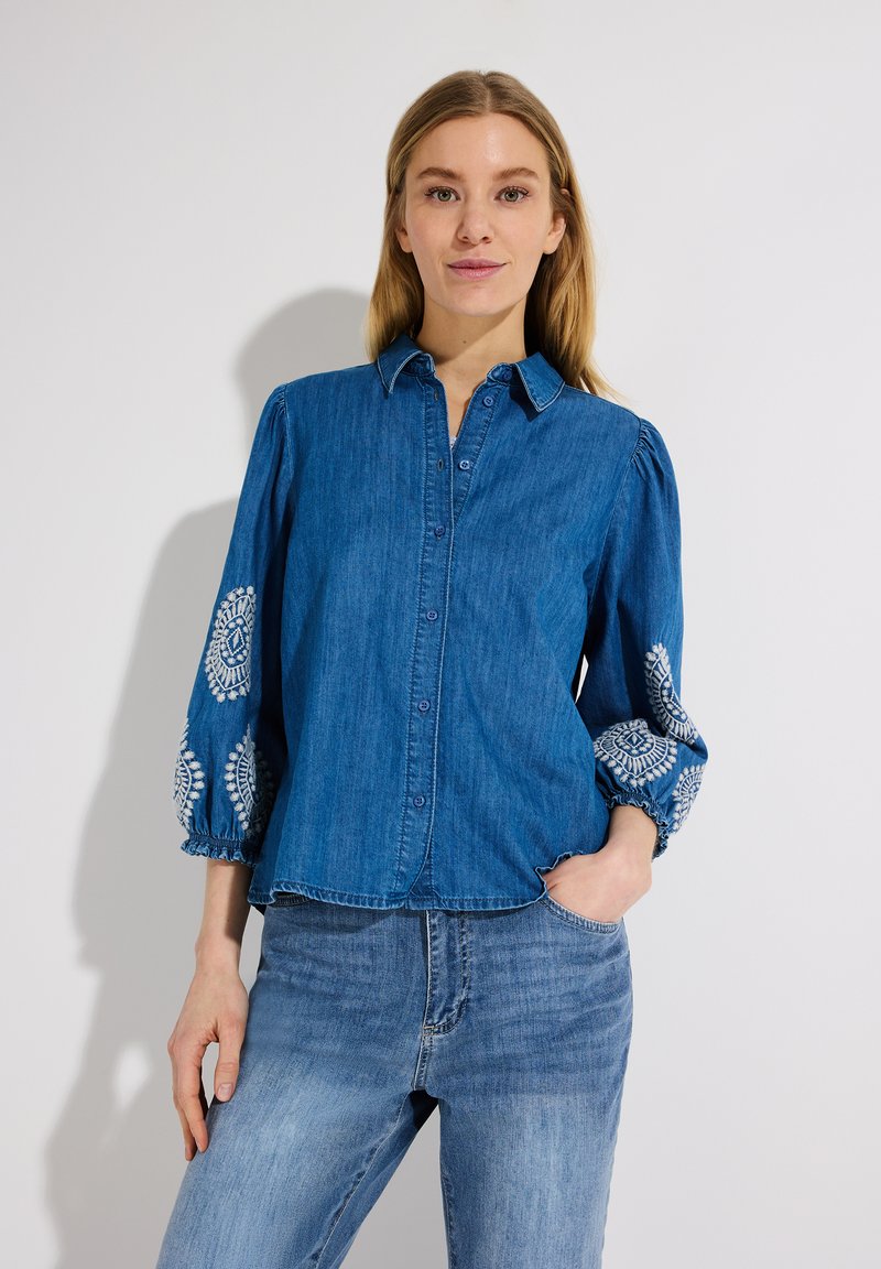 Donna che indossa una camicia blu di jeans con bottoni e motivi ricamati bianchi sulle maniche, abbinata a jeans di denim azzurro chiaro, su sfondo neutro.