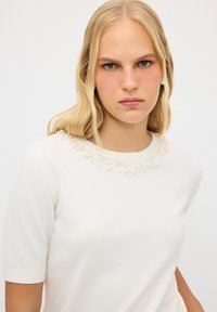 Vit kortärmad tröja med rundad halsringning dekorerad med texturerade konstgjorda pärlor runt kragen. Slätt stickat tyg.