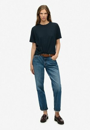 T-shirt en coton noir à manches courtes, associé à un jean droit bleu clair et des mocassins marron foncé, se tenant contre un fond blanc.