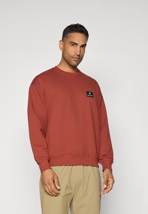 Sweatshirt couleur rouille avec un col rond, des manches longues, et un patch rectangulaire noir affichant un logo sur le devant.