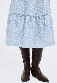 Φόρεμα ανοιχτού μπλε χρώματος με λευκό πουά υφή, συνδυασμένο με δερμάτινες μπότες μέχρι το γόνατο σε σκούρο καφέ χρώμα με διακοσμητικές ραφές.