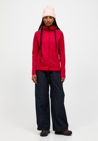 Rode zip-up jas van gladde stof, gecombineerd met losse marineblauwe broek met een gestructureerde afwerking. Lichtroze gebreide beanie op het hoofd.