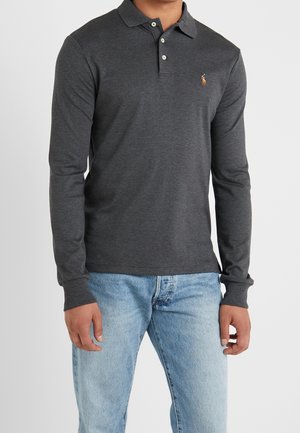 Homme portant un polo à manches longues gris foncé avec un logo brodé et un jean bleu clair, debout devant un fond uni.