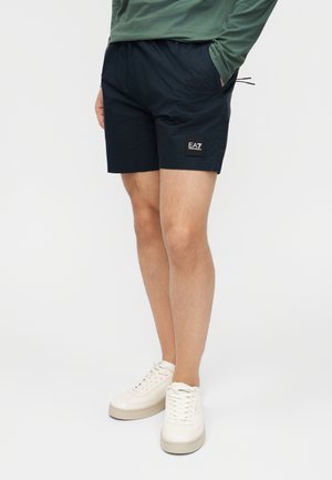 Άνδρας που φοράει μαύρα σορτς EA7, πράσινο μακρυμάνικο πουκάμισο και λευκά αθλητικά παπούτσια, στέκεται με τα χέρια στις τσέπες σε λευκό φόντο.