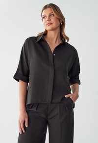Camicia nera a maniche corte con bottoni realizzata in tessuto morbido. Caratterizzata da un colletto, maniche rimboccate e una vestibilità ampia. Design minimalista senza fantasie.