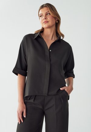 Black short-sleeve button-up shirt made of smooth fabric. Features a collar, rolled sleeves, and a loose fit. Minimalistic design with no patterns.