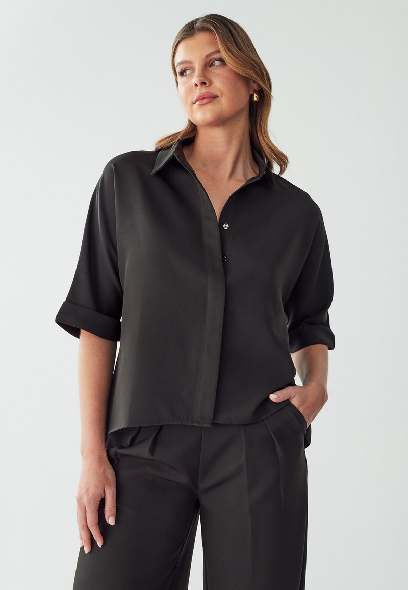 Camisa negra de botones de manga corta hecha de tela suave. Presenta un cuello, mangas enrolladas y un ajuste holgado. Diseño minimalista sin patrones.