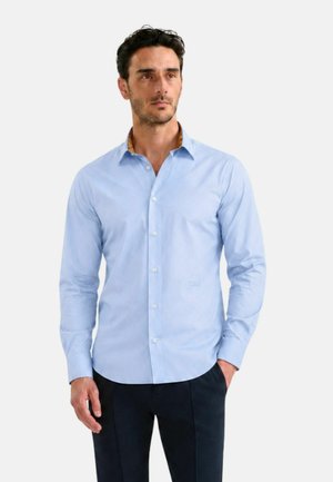 Uomo con capelli scuri che indossa una camicia azzurro chiaro con bottoni e pantaloni scuri, con una mano in tasca, in piedi davanti a uno sfondo semplice.