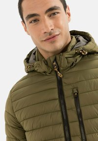 Olivgrüne Pufferjacke mit horizontalem Steppmuster, Reißverschluss vorne, verstellbarem Kapuzenrand und orangefarbenen Hardware-Elementen.