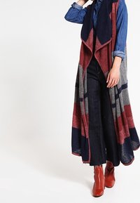 Person som bär en lång ärmlös kofta i röd, marinblå och grå över en uppvikt denimskjorta, mörka vida jeans och röda ankelskor.