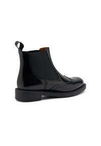Botín Chelsea de cuero negro con paneles elásticos en los laterales, detalles de brogue en la punta y un tacón de goma bajo. Textura lisa con un acabado brillante.