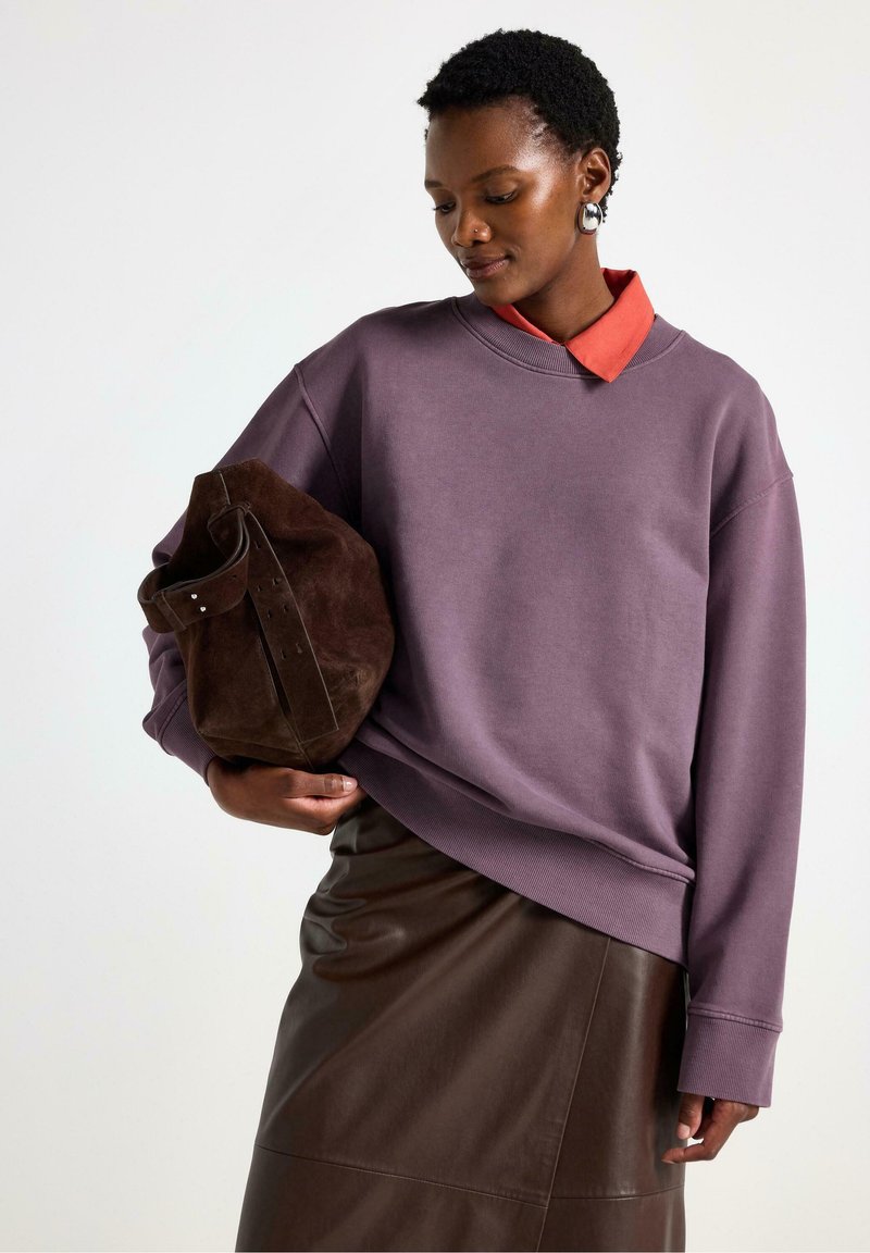 Sweatshirt viola oversize con scollo a giro sopra una camicia rossa con colletto, abbinato a una borsa in suede marrone e una gonna in pelle marrone.