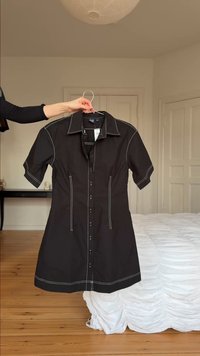 Χέρι που κρατάει μαύρο φόρεμα με κοντά μανίκια, κουμπιά και λευκές ραφές, κρεμασμένο σε κρεμάστρα σε υπνοδωμάτιο με ξύλινο πάτωμα και λευκό κρεβάτι.