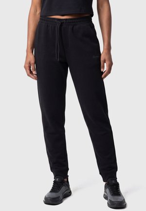 Femme portant un pantalon de jogging noir avec une taille à cordon et des chaussures de sport noires et grises, debout devant un fond clair uni.