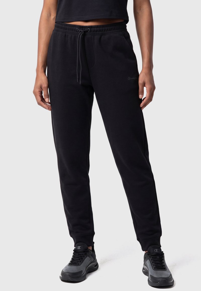 Femme portant un pantalon de jogging noir avec une taille à cordon et des chaussures de sport noires et grises, debout devant un fond clair uni.