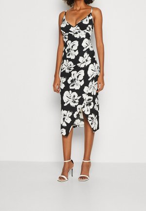 Robe midi noire avec un grand imprimé floral blanc, bretelles spaghetti, fente devant, portée avec des sandales blanches à talons hauts sur fond uni.
