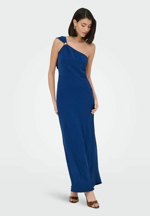 Kvinde iført en lang, blå kjole med én skulder og en guldlås, iført brune hæle, stående og kigger ned mod sin venstre side.