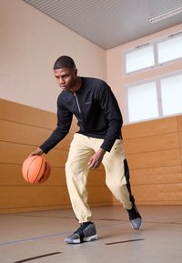 Sort lynlås langærmet top, lysegule joggers med sorte accenter, grå sportssko, orange basketball, indendørs sportsbane i baggrunden.