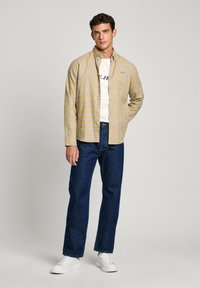 Camisa de franela a cuadros amarilla y azul, abotonada, sobre una camiseta blanca con estampado negro; combinada con jeans azul oscuro y zapatillas blancas.
