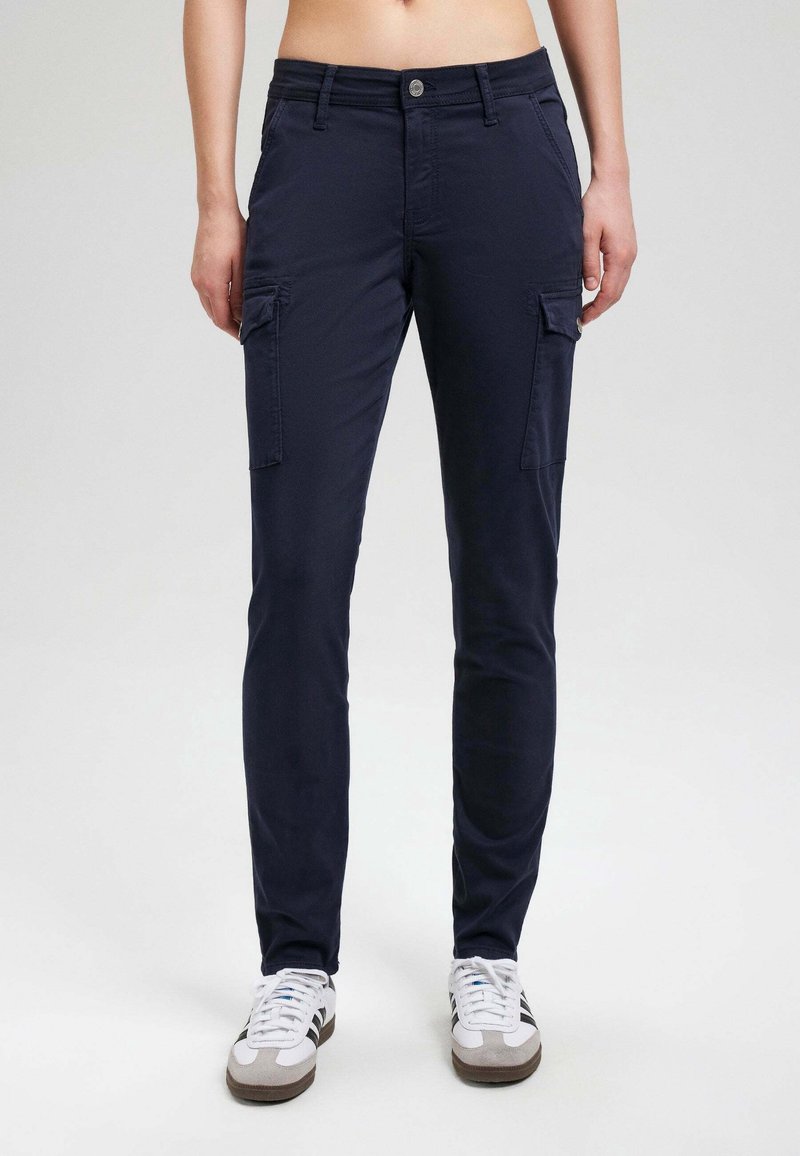 Personne portant un pantalon cargo bleu marine avec poches à rabat à l'avant et des baskets blanches, debout devant un fond clair uni.