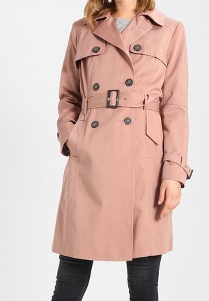 Femme portant un trench-coat mi-long rose poudré à double boutonnage, taille ceinturée et poches latérales, associé à un pantalon foncé.