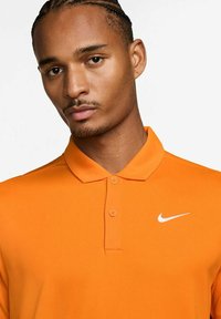 Narancssárga póló, struktúrált gallérral, két gombbal és egy kis fehér Nike logóval a bal mellen. Simább, textúrált anyag.