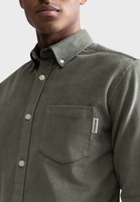 Homme portant une chemise boutonnée texturée vert olive avec poche poitrine et petite étiquette "Redefined Rebel" sur le bord de la poche.
