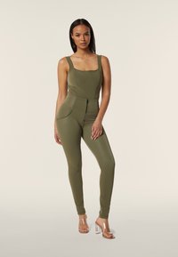 Jumpsuit verde oliva con scollatura quadrata, design aderente, vita alta e zip frontale. Abbinato a sandali con tacco trasparente.