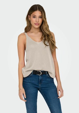 Young woman with long wavy hair wearing beige sleeveless top, blue jeans, black belt, gold hoop earrings, necklace, and bracelet, standing against white background.