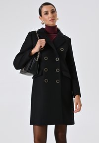 Cappotto nero doppiopetto con dettagli in tonalità dorata, colletto a punte e tasche laterali. Indossato sopra un dolcevita bordeaux. Tessuto strutturato.