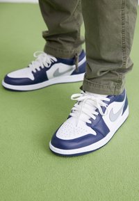 Person i vita och marinblå Nike-sneakers med grå swoosh på en grön matta, byxor upptäckta strax ovanför anklarna.