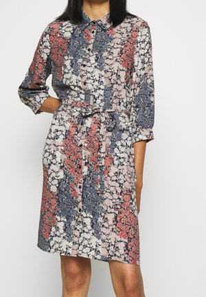 Vrouw draagt een knielange bloemenoverhemdjurk met blauwe, witte en rode bloemen, driekwart mouwen en een ceintuur in de taille.