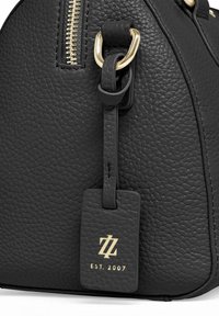 Gros plan d’un sac en cuir noir texturé avec une fermeture éclair dorée et une étiquette logo dorée portant l’inscription "EST. 2007", suspendue à un anneau doré.