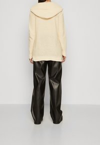 Osoba oblečená v krémové pletené mikině s kapucí a volných černých kožených kalhotách, stojící zády k jednoduchému světle šedému pozadí.