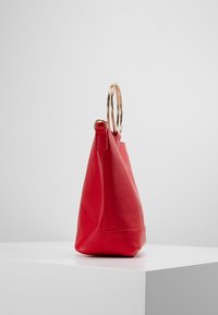 Rød lædertaske med trekantet form, med to guldfarvede metalringe som håndtag og glat overflade med flad bund.