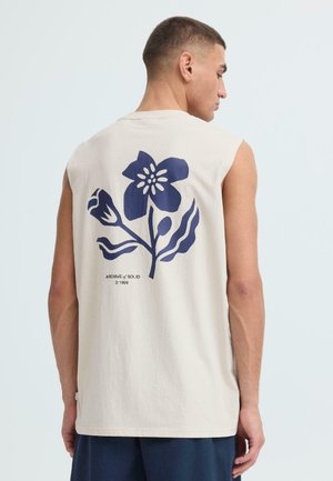 Jonge man die opzij kijkt, draagt een mouwloos beige shirt met een grote donkerblauwe bloemenprint op de achterkant en marineblauwe korte broek.