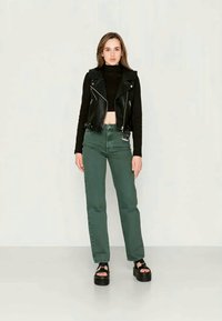 Jovem mulher de pé, vestindo um colete de couro preto sem mangas, uma blusa crop de mangas longas preta, jeans verdes de cintura alta e sandálias de plataforma pretas.