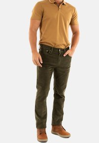 Camisa polo marrón, pantalones verde oliva y botas de tobillo marrones. Corte slim con detalles en los bolsillos y una textura suave. Esquema de colores simple, look casual.