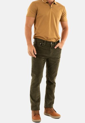 Camisa polo marrón, pantalones verde oliva y botas de tobillo marrones. Corte slim con detalles en los bolsillos y una textura suave. Esquema de colores simple, look casual.