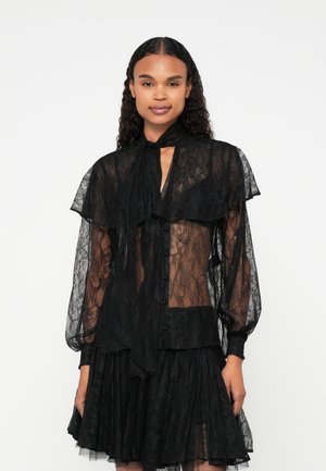 Musta pitsipusero, jossa on läpinäkyvä kangas, korkea kaulus nauhalla, pitkät puffihihat ja nappikiinnitys. Kerroksellinen muotoilu, jossa on tekstuuroituja yksityiskohtia.