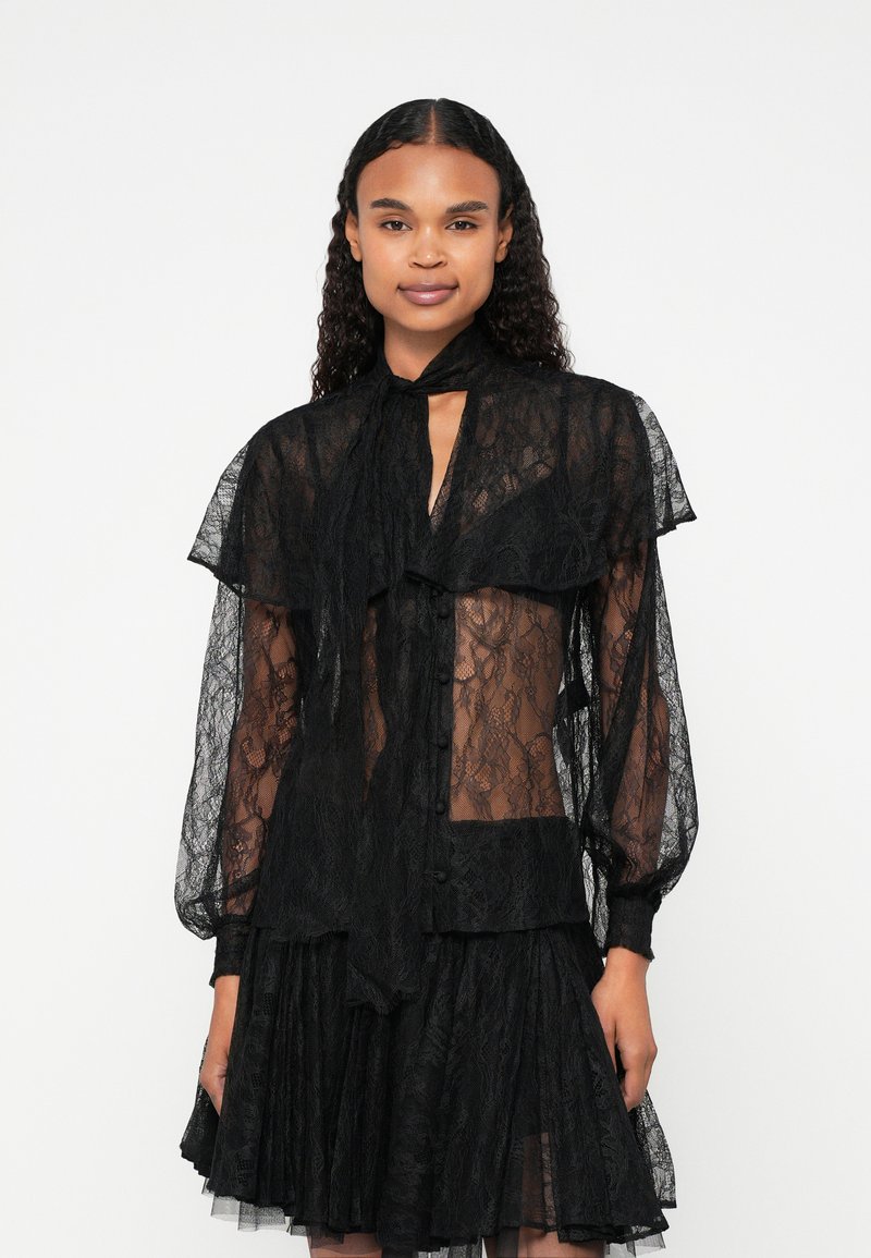 Zwarte kantblouse van doorschijnende stof, hoge hals met strik, lange pofmouwen en knoopsluiting. Gelaagd ontwerp met textuuraccenten.