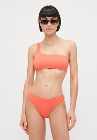 Set de bikini coral cu un umăr, realizat dintr-un material texturat; chilot cu talie joasă asortat. Ochelari de soare cu model țestoasă.